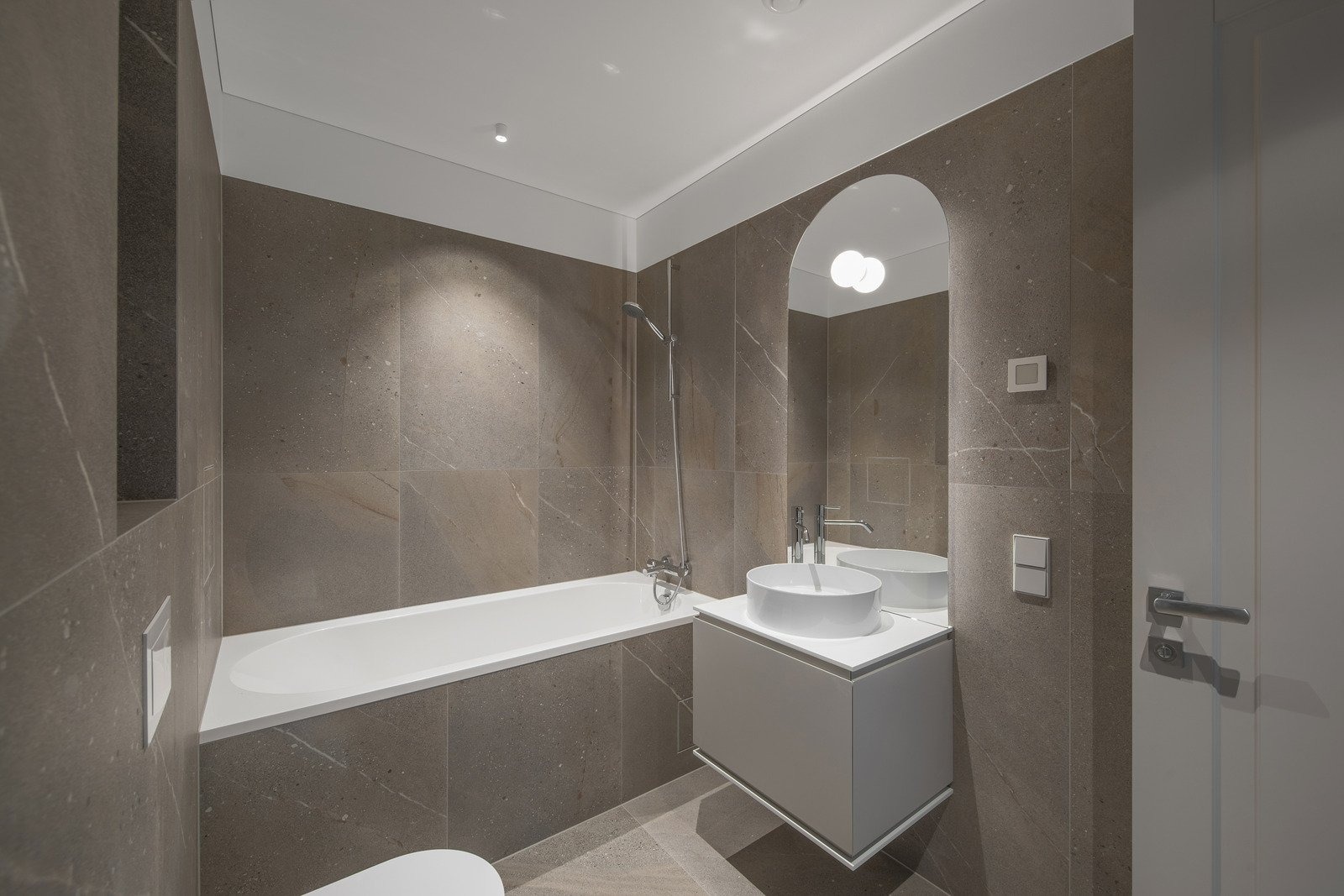 Nowoczesna łazienka wykończona płytkami imitującymi kamień, z białą wanną, umywalką nablatową i lustrem w kształcie łuku.