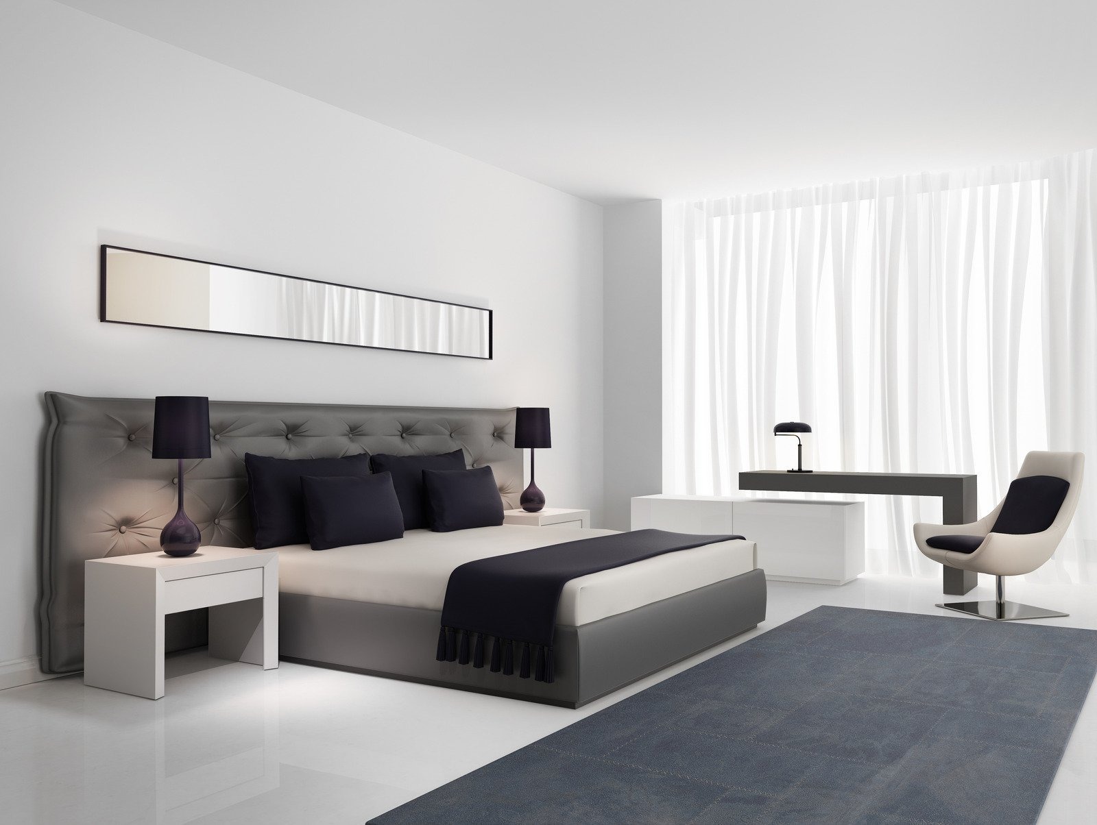 Minimalistyczna sypialnia z szarym łóżkiem z pikowanym zagłówkiem, ciemnoniebieskimi poduszkami i narzutą z frędzlami, białe szafki nocne z lampkami, długie lustro nad łóżkiem, biurko z krzesłem...