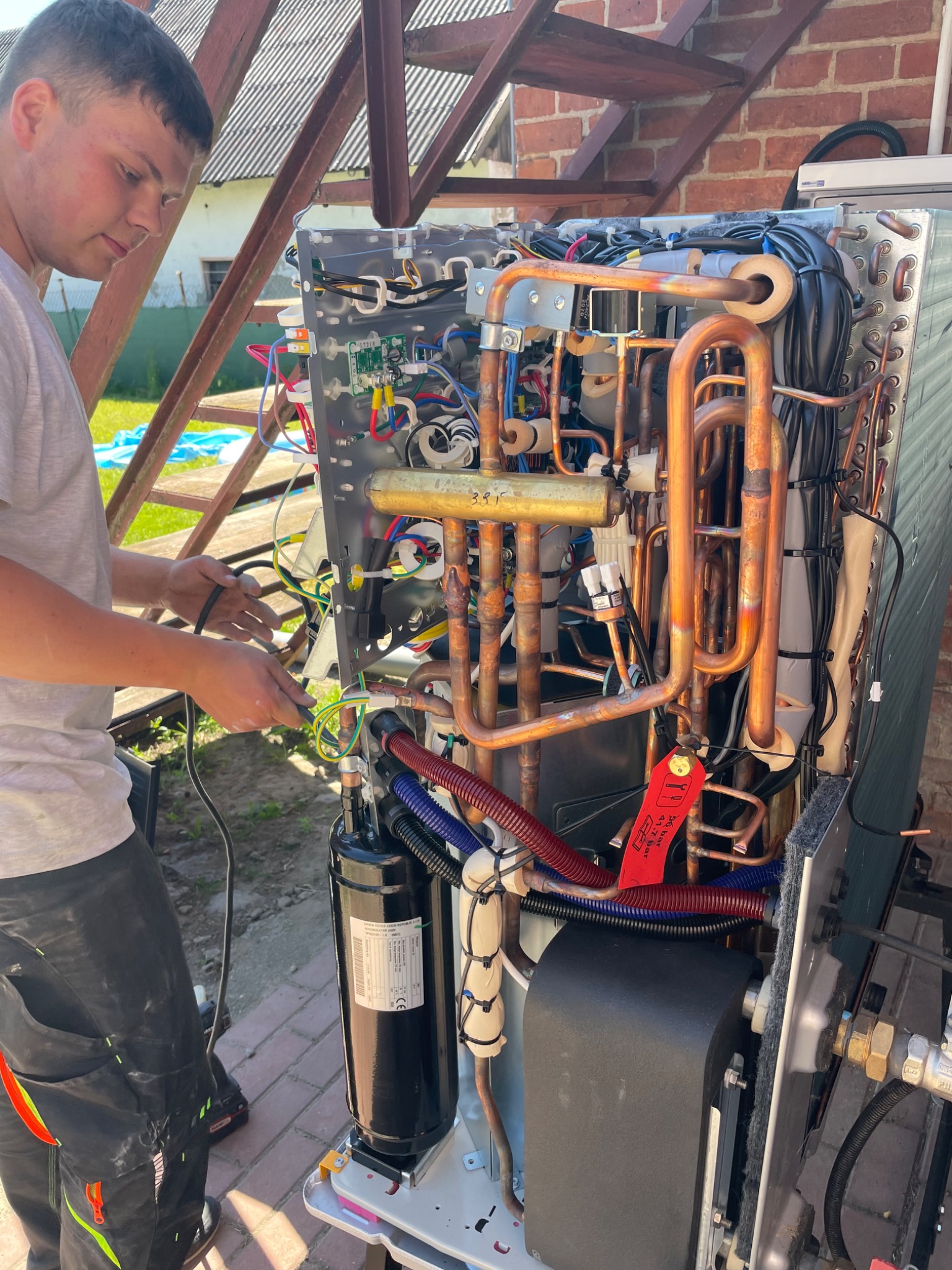 Młody mężczyzna podłącza przewody wewnątrz otwartej obudowy pompy ciepła, widoczne miedziane rurki i elementy elektroniczne, w tle ceglany budynek i drewniana konstrukcja.