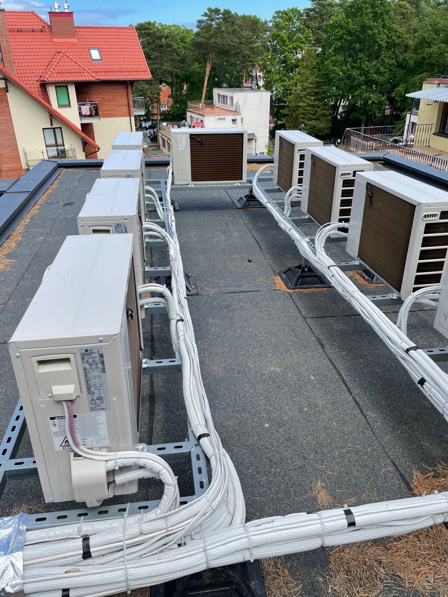 Rząd jednostek zewnętrznych klimatyzacji LG na dachu pokrytym papą, z widocznymi przewodami w izolacji, zamocowanymi do metalowej konstrukcji wsporczej.