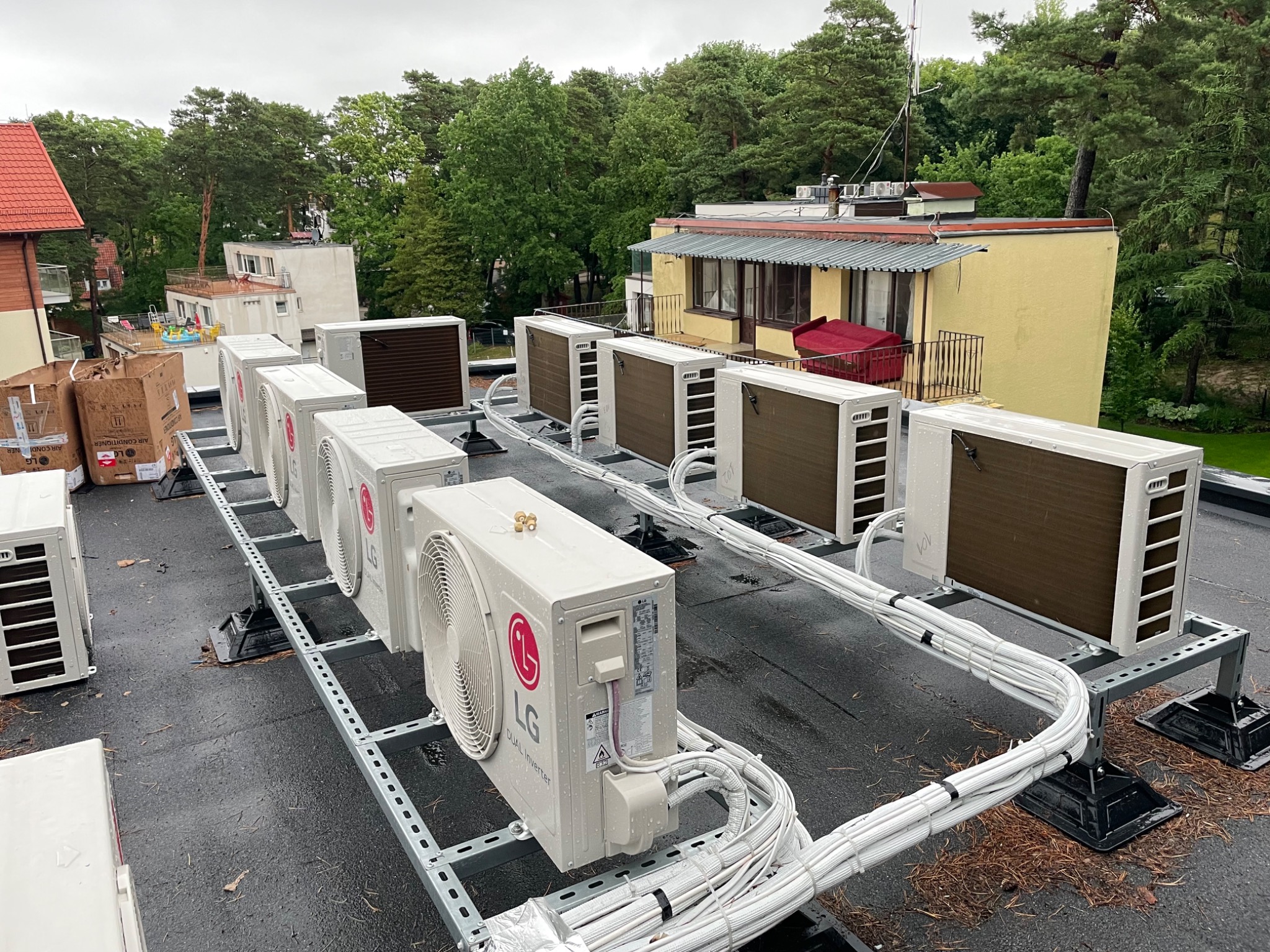 Rząd zewnętrznych jednostek klimatyzacji LG Dual Inverter zamontowanych na metalowej konstrukcji na dachu budynku, widoczne białe kable instalacyjne. W tle budynki i drzewa.