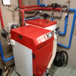 Nowoczesny piec z podajnikiem, z czerwono-białą obudową, widoczne instalacje grzewcze w kolorze czerwonym i niebieskim, szare płytki na ścianach i podłodze.