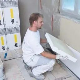 Pracownik w białych ubraniach montuje białe płyty izolacyjne STO na ścianie wewnętrznej, widoczny klej na ścianie, wiadro z klejem i paczki płyt izolacyjnych STO w tle.