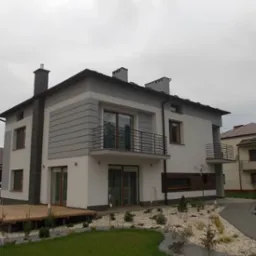 Nowoczesny dom dwukondygnacyjny z balkonami, wykończony w bieli i szarości, z drewnianym tarasem i kamienną ścieżką w ogrodzie.