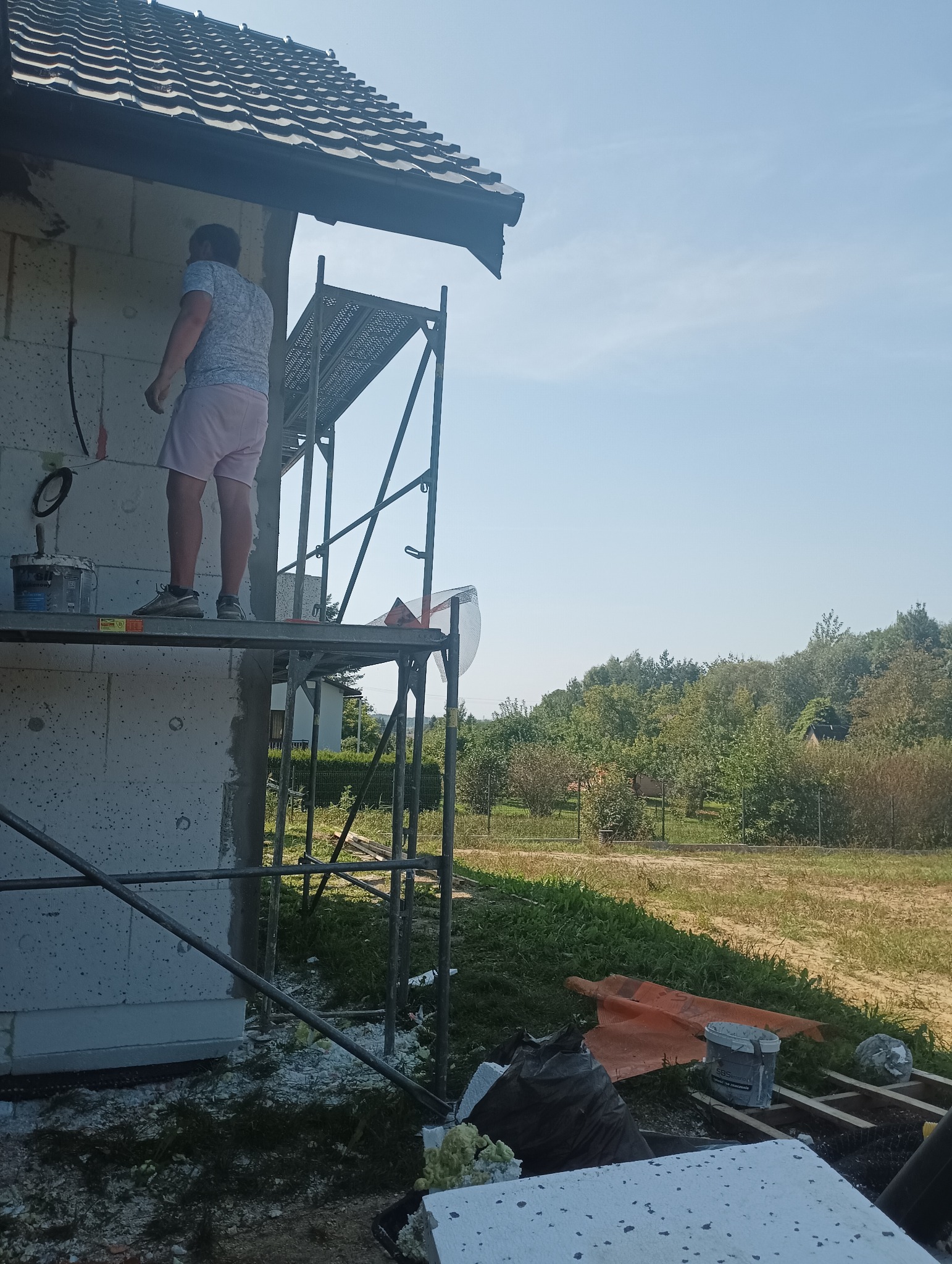 Pracownik na rusztowaniu przy ocieplaniu ściany domu z betonu komórkowego, widoczne puszki z klejem, fragment dachu, w tle zieleń.