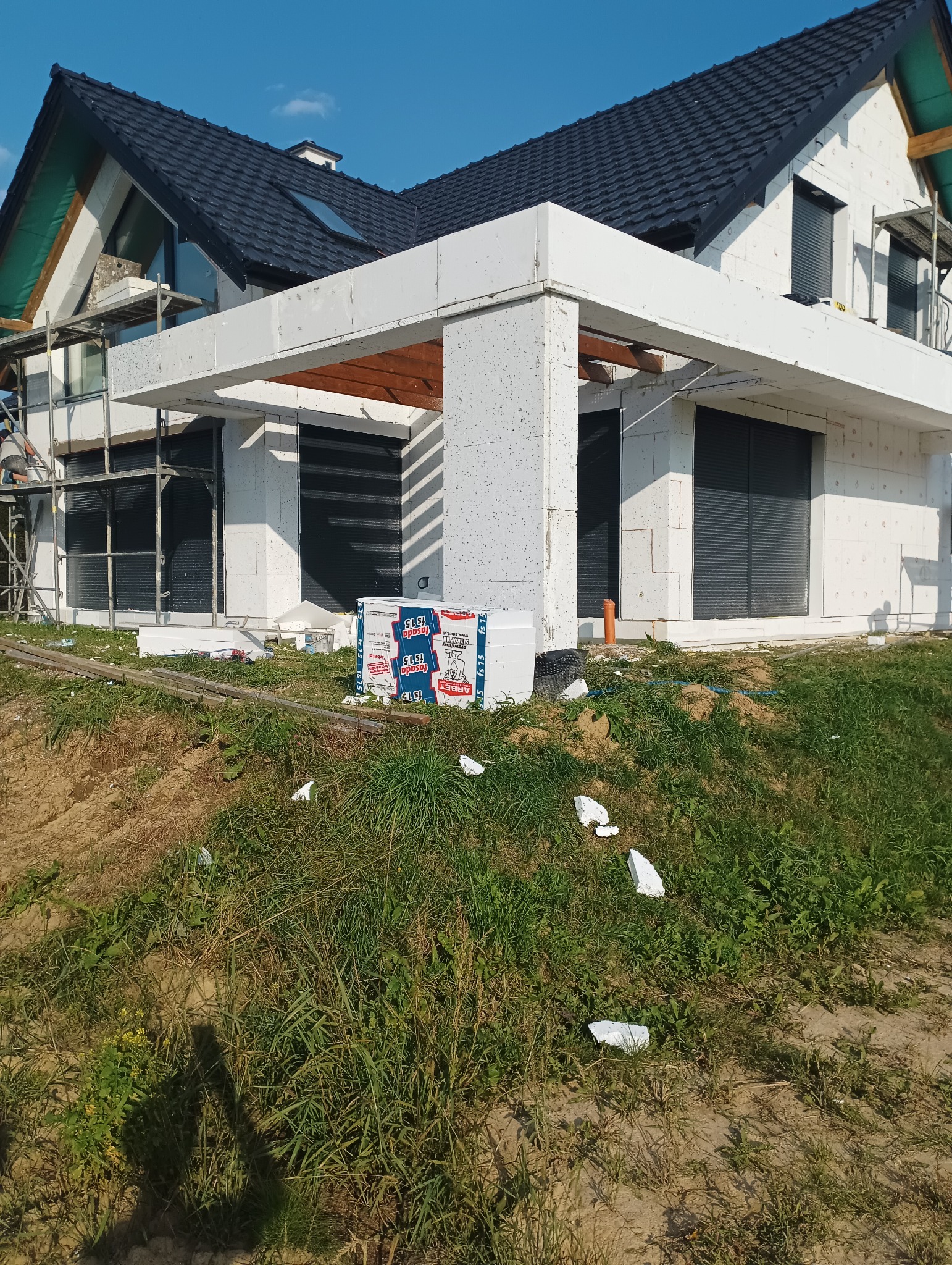 Niedokończony dom z białych bloczków, widoczne rusztowania, okna z roletami zewnętrznymi, paczki styropianu na trawie.