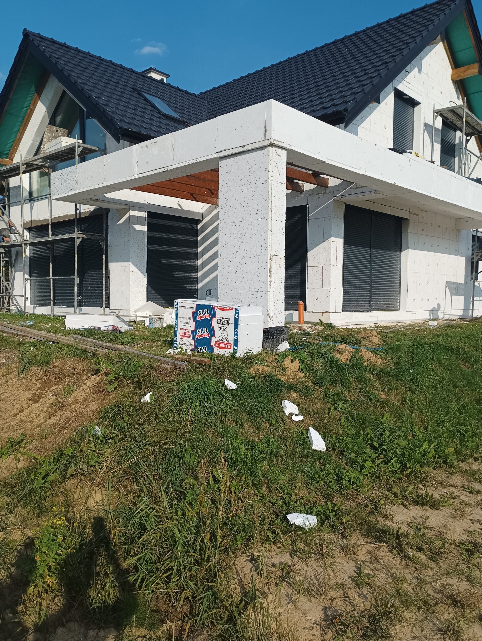 Surowy stan budynku z białego betonu komórkowego, okna z roletami zewnętrznymi, w tle rusztowania i dach z czarnej dachówki, na pierwszym planie paczki materiałów budowlanych i ścinki styropianu...