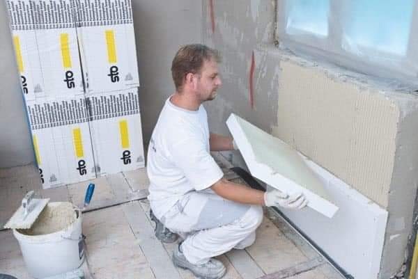 Pracownik w białych ubraniach montuje białe płyty izolacyjne STO na ścianie wewnętrznej, widoczny klej na ścianie, wiadro z klejem i paczki płyt izolacyjnych STO w tle.