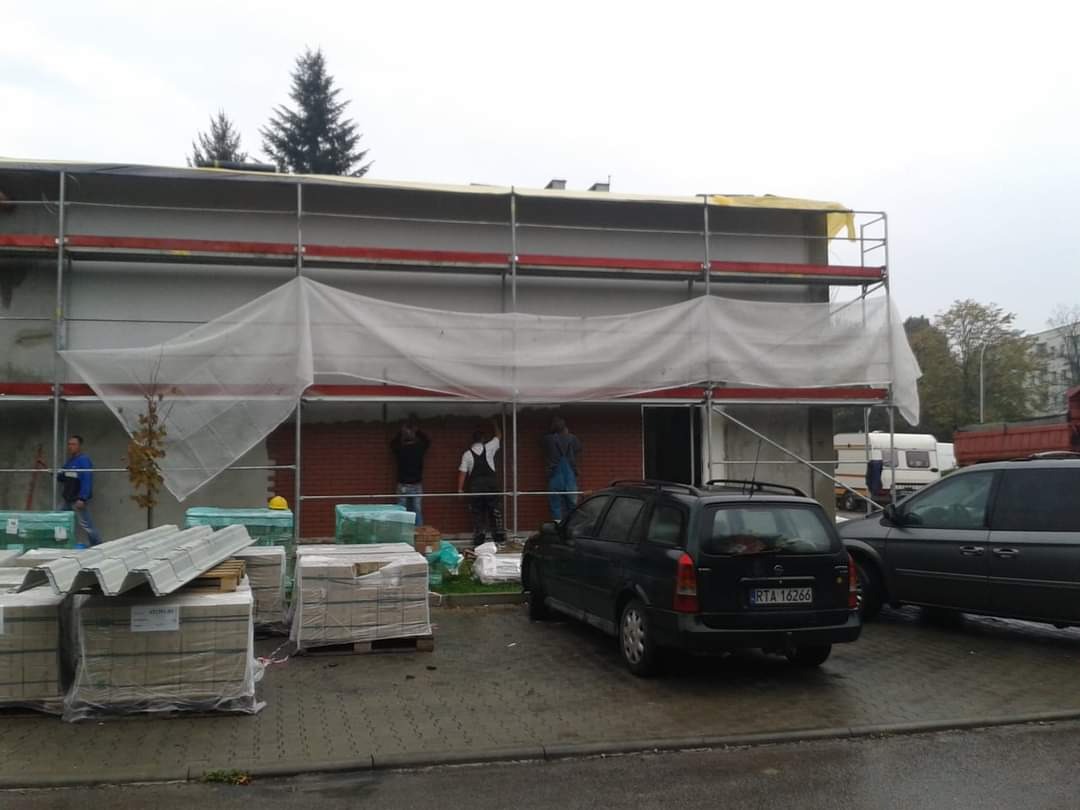 Prace murarskie na zewnątrz budynku w trakcie realizacji, widoczne rusztowania, palety z cegłami, pracownicy w trakcie murowania ściany z czerwonej cegły, zaparkowane samochody.