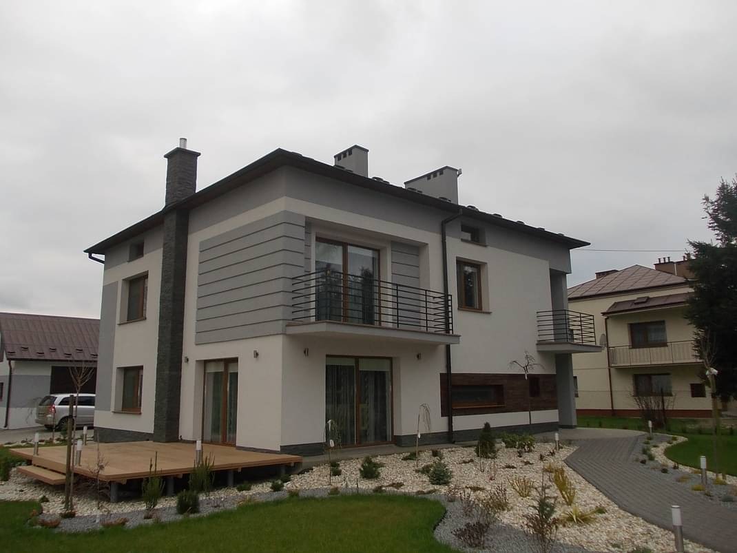 Nowoczesny dom dwukondygnacyjny z balkonami, wykończony w bieli i szarości, z drewnianym tarasem i kamienną ścieżką w ogrodzie.