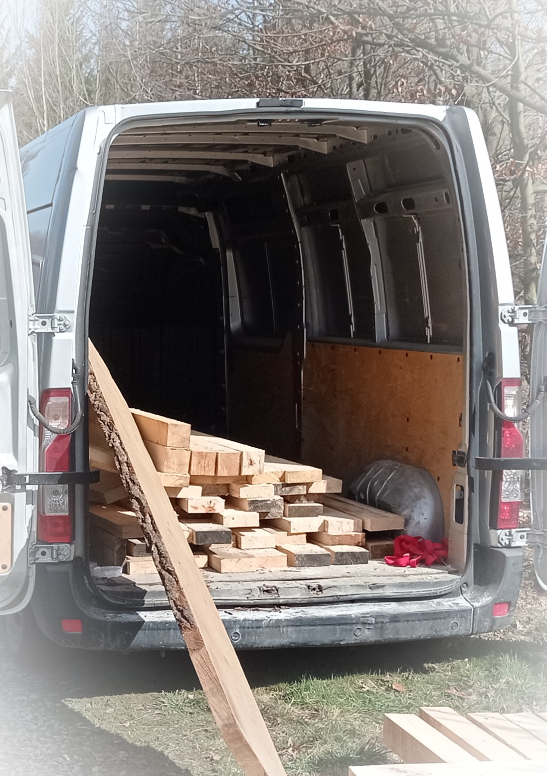 Wnętrze białego busa z otwartymi drzwiami załadowane stertą drewnianych desek i kantówek o jasnym odcieniu, niektóre z korą, na tle drzew bez liści.