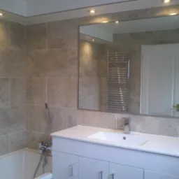 Wykończona łazienka z beżowymi płytkami, dużym lustrem i białą szafką pod umywalką. Widoczne oświetlenie punktowe i grzejnik drabinkowy w odbiciu lustra.