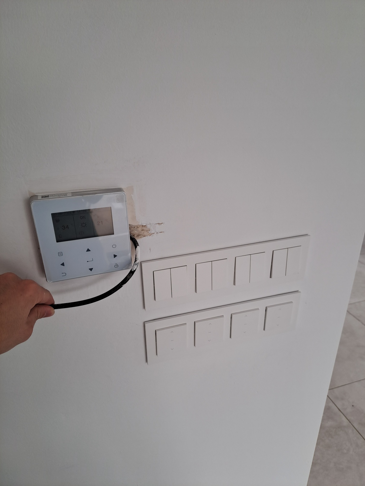 Biały panel sterowania ogrzewaniem podłogowym z wyświetlaczem, zamontowany na ścianie obok zestawu włączników światła, widoczne ślady po montażu, w dłoni trzymany kabel.