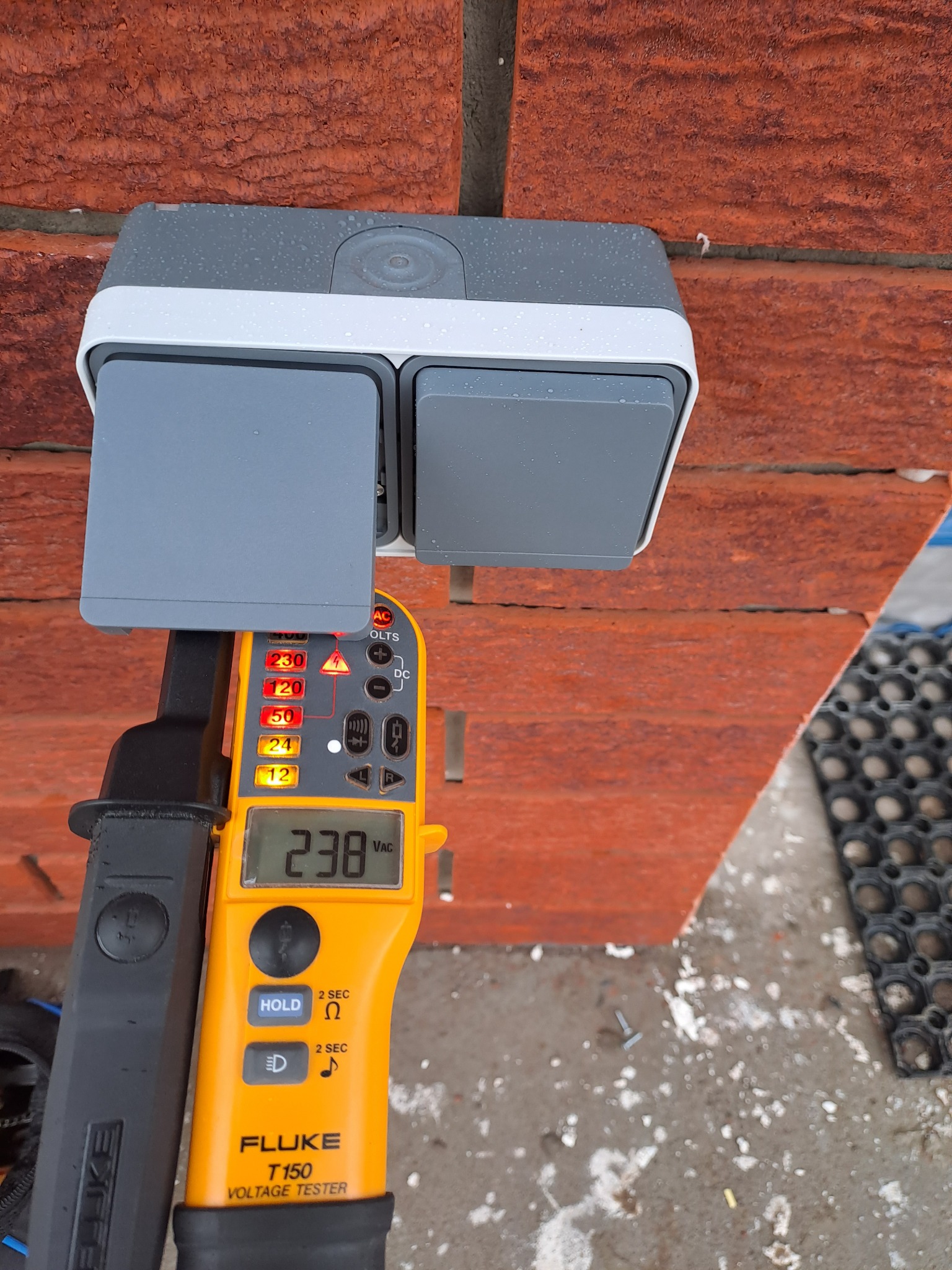 Szary podwójny łącznik natynkowy na ceglanej ścianie, sprawdzany miernikiem napięcia Fluke T150, wskazującym 238 VAC.