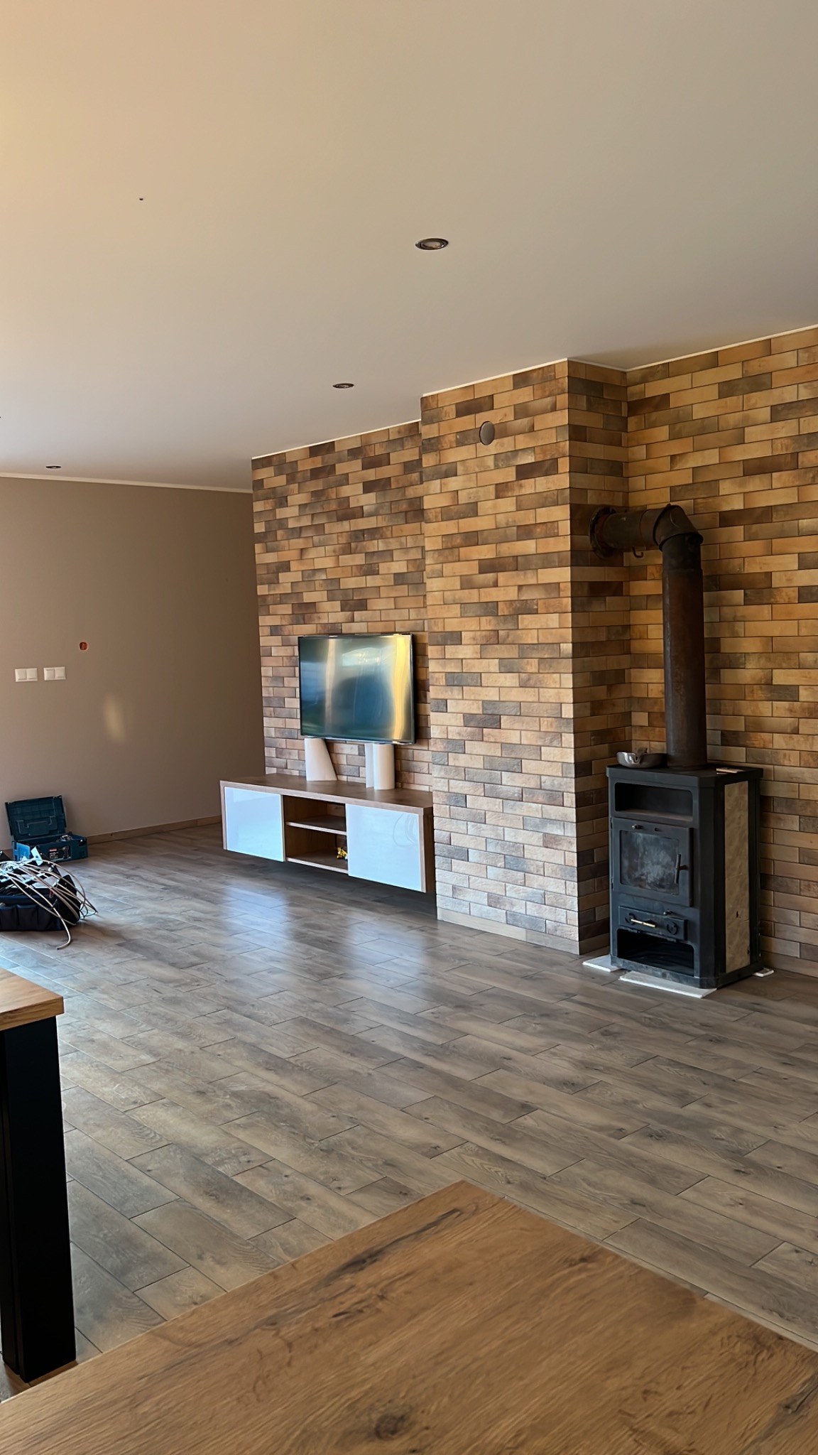 Salon z ceglaną ścianą, kominkiem typu koza, drewnianą podłogą i szafką RTV pod telewizorem, częściowo widoczny blat stołu na pierwszym planie.