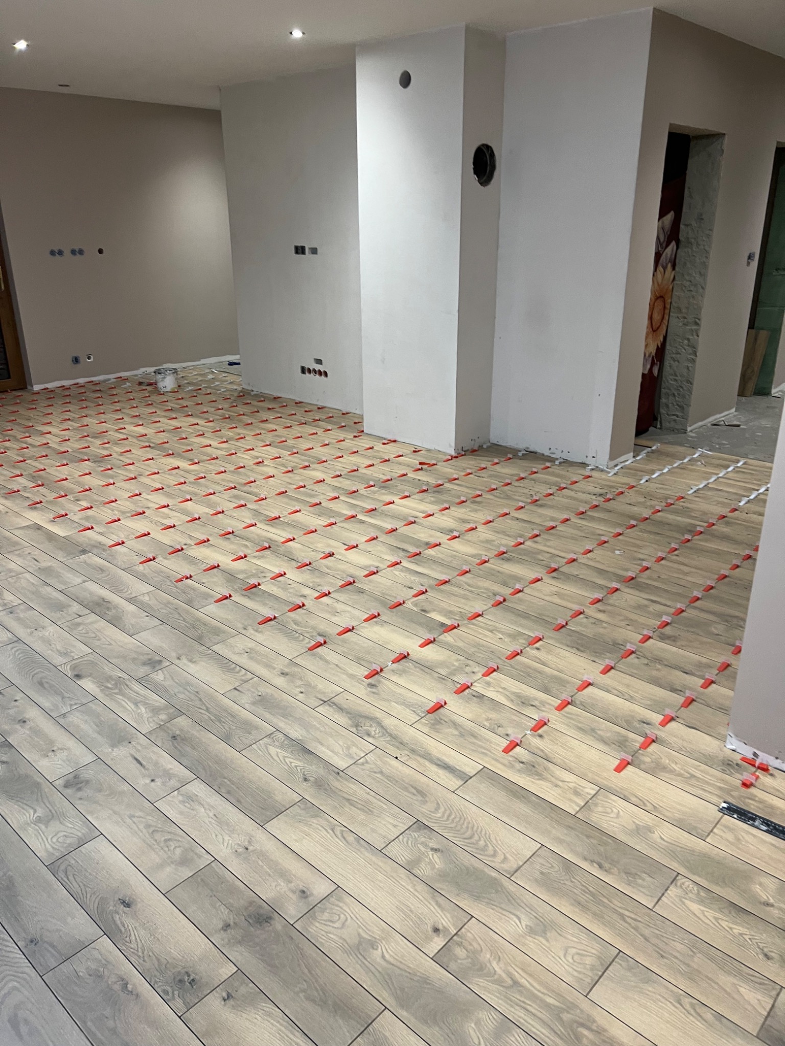 Układanie podłogi z płytek imitujących drewno z użyciem czerwonych krzyżyków dystansowych; widoczne przygotowane, surowe ściany i instalacje elektryczne.