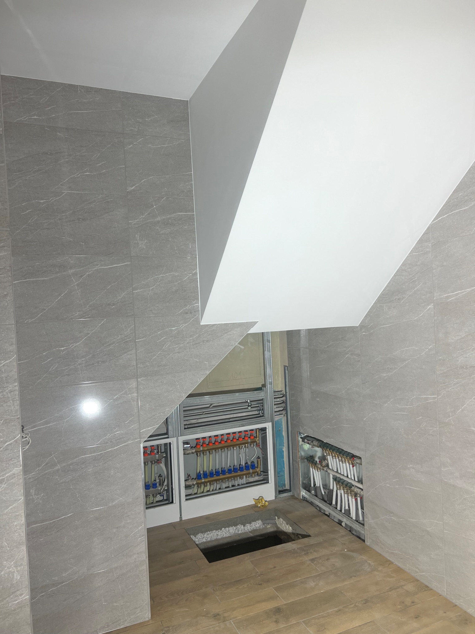 Łazienka w trakcie remontu: szare płytki imitujące marmur na ścianach, częściowo położona drewnopodobna podłoga, widoczne instalacje hydrauliczne i elektryczne w ścianach, sufit podwieszany...