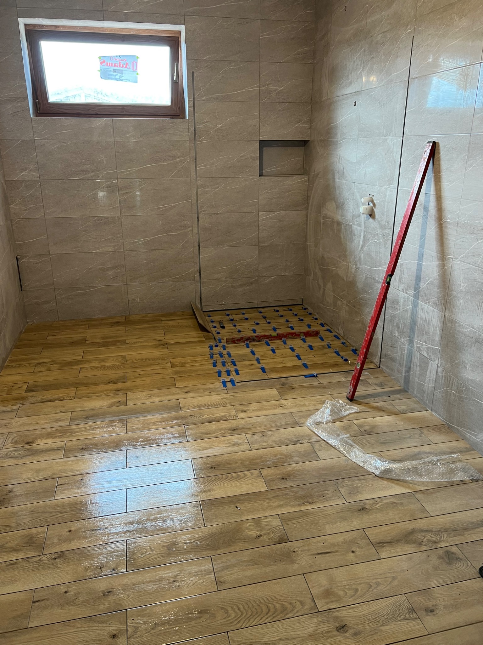 Łazienka w trakcie układania płytek podłogowych imitujących drewno, z widocznym poziomem i elementami dystansowymi, ściany wyłożone płytkami w odcieniach beżu z szarymi żyłkami.