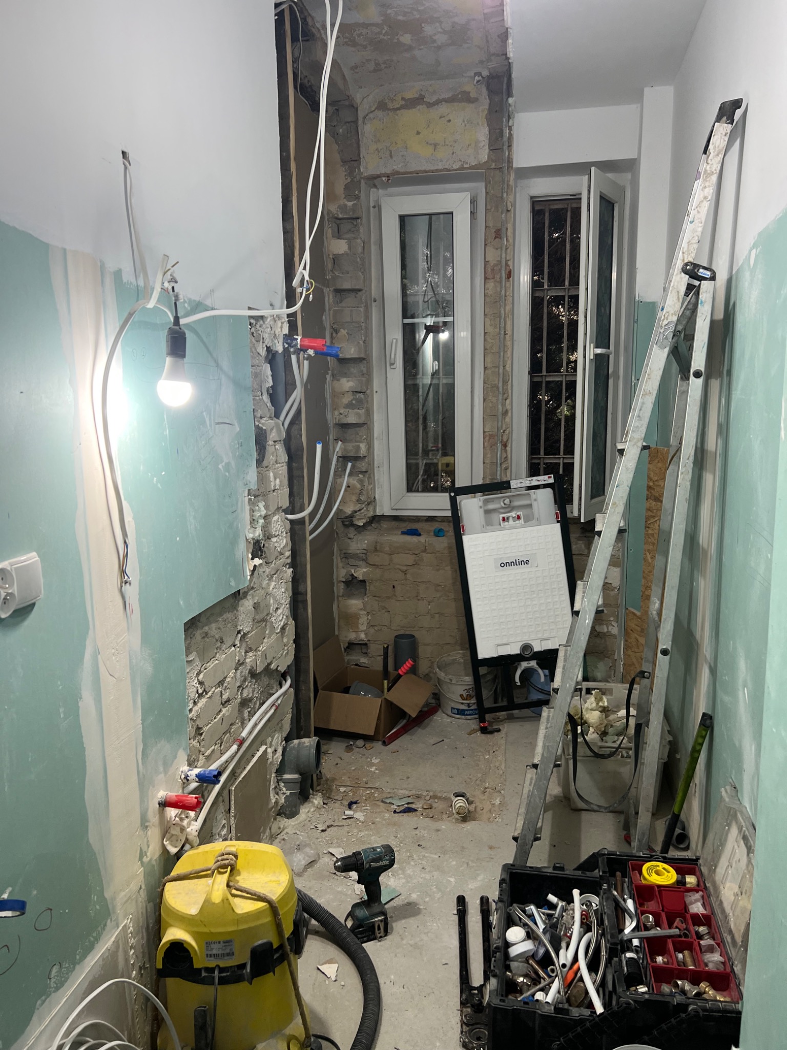 Remont łazienki w trakcie: widoczne odsłonięte rury, stelaż podtynkowy WC marki Onnline, drabina, narzędzia glazurnicze oraz odkurzacz budowlany