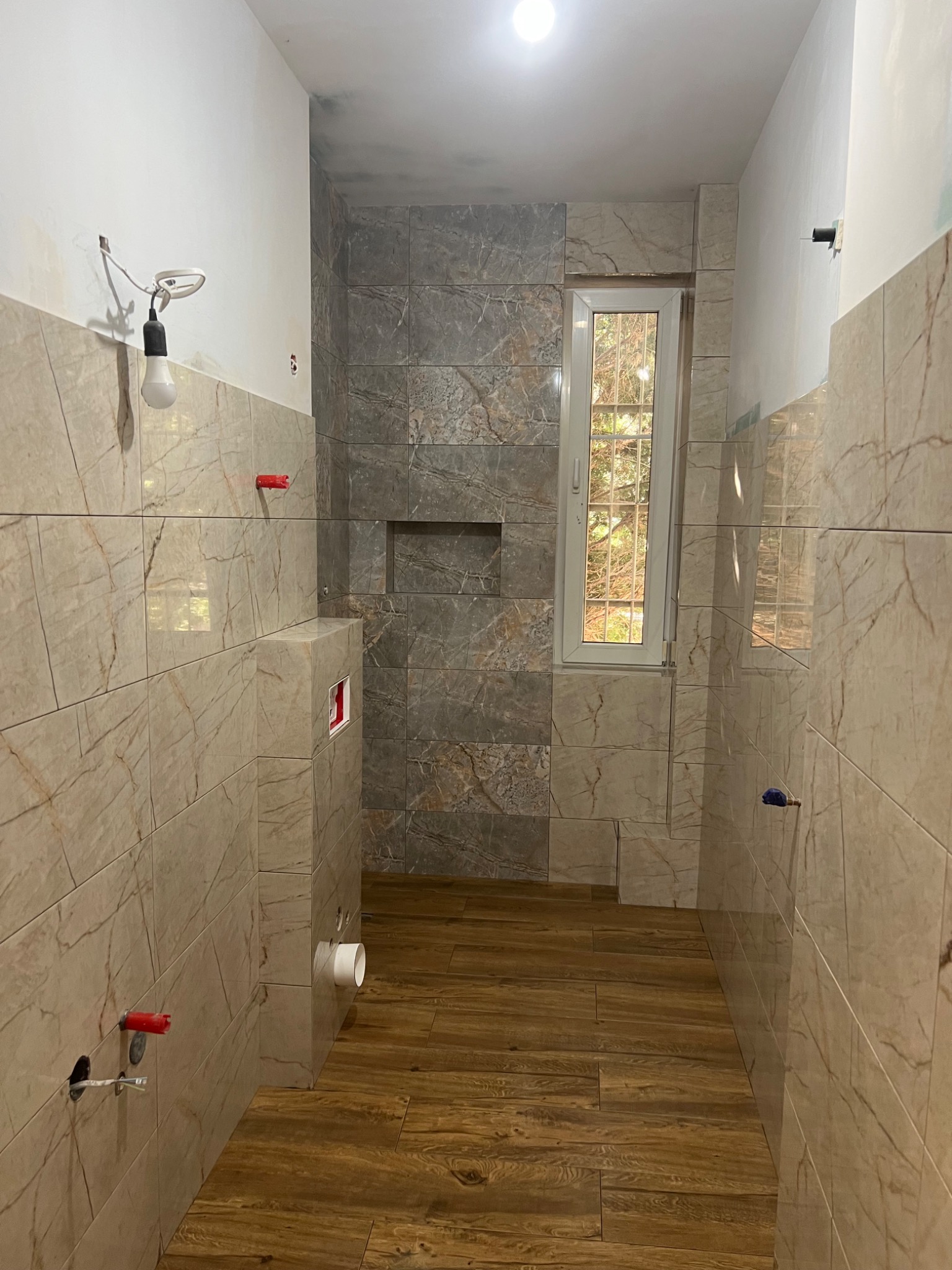 Wnętrze łazienki w trakcie remontu w Krajenka, widoczne płytki imitujące marmur na ścianach i drewnopodobne na podłodze, przygotowane instalacje wodno-kanalizacyjne oraz okno z widokiem na zieleń.