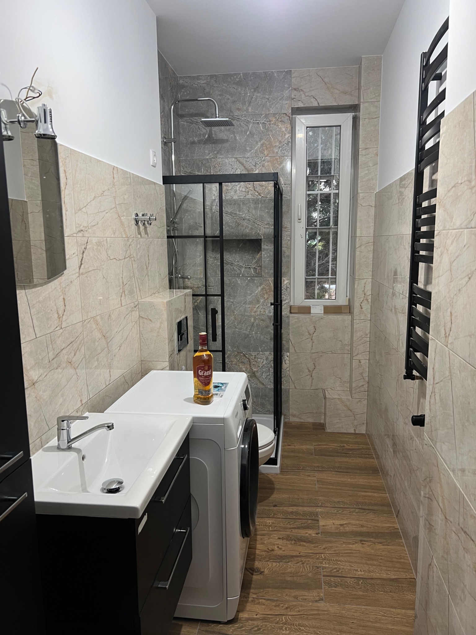 Ukończona łazienka z beżowymi i szarymi płytkami imitującymi marmur, kabina prysznicowa z czarną ramą, pralka z butelką whisky na górze, czarna szafka pod umywalką i grzejnik drabinkowy.