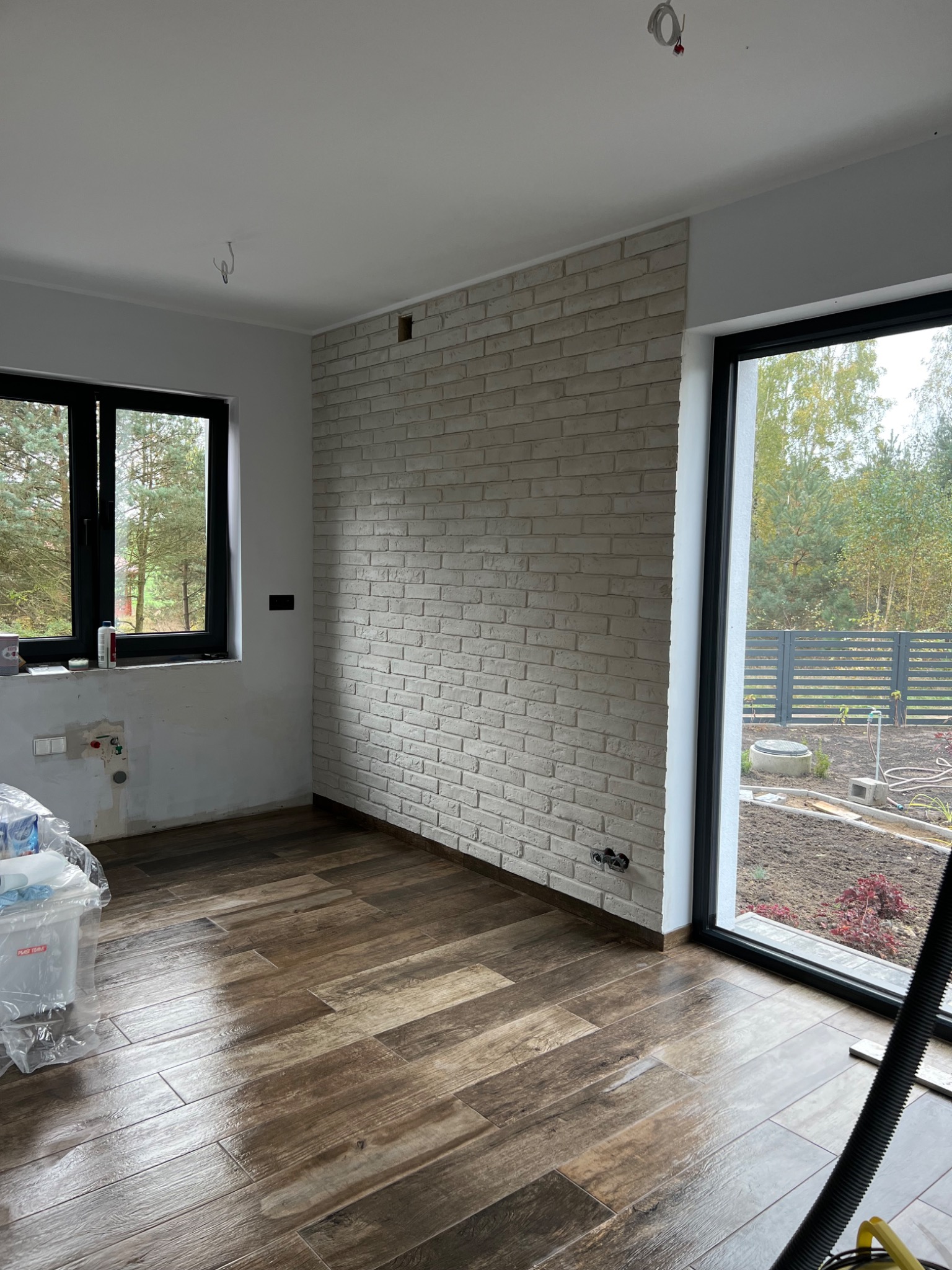 Wnętrze w trakcie prac: ściana wyłożona białą cegłą, brązowa podłoga imitująca drewno, czarne ramy okienne, widoczne wyprowadzenia instalacji elektrycznej i hydraulicznej, plastikowe wiadra...