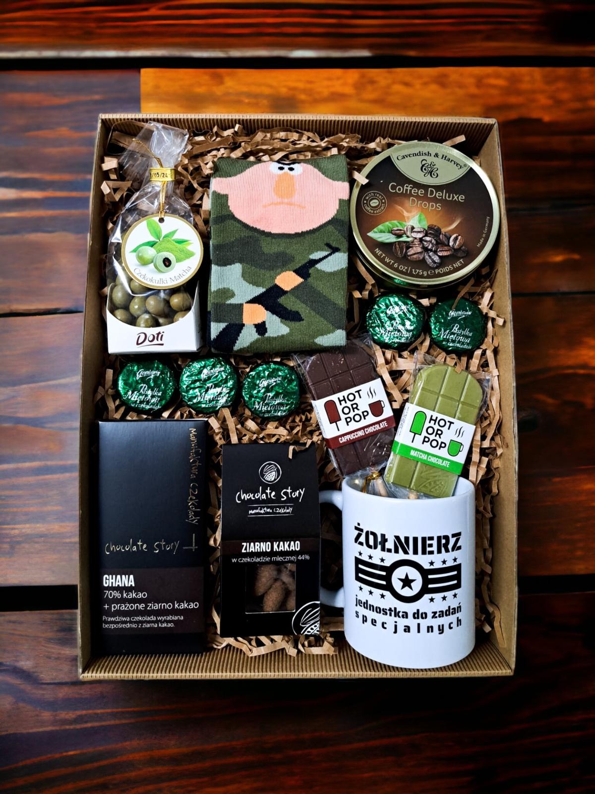 Prezentowy zestaw w kartonowym pudełku wypełnionym papierowym siankiem, zawierający słodycze, kawę w puszce, skarpetki z motywem żołnierza i kubek z napisem 'Żołnierz jednostka do zadań specjalnych'.
