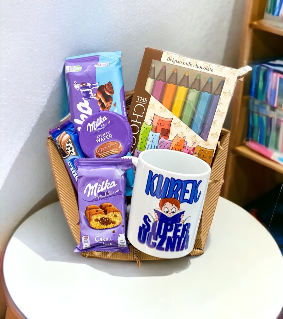 Wiklinowy kosz wypełniony słodyczami marki Milka, batonami Oreo, czekoladą w kształcie kredek oraz kubkiem z napisem 'Kurek Super Ucznia', na białym stole.