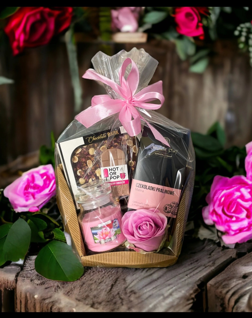 Elegancki upominek w celofanie z różową wstążką, zawierający czekolady, świecę zapachową i dekoracyjną różę, otoczony kwiatami na rustykalnym tle.