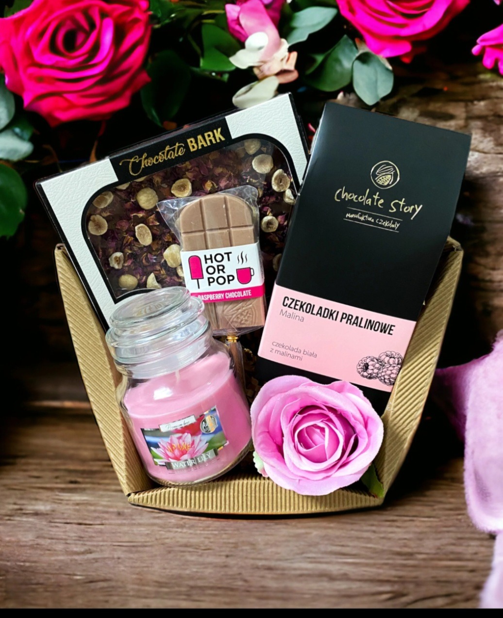 Kosz prezentowy z czekoladkami Chocolate Story, tabliczką Chocolate Bark z orzechami i różami, czekoladą malinową Hot or Pop oraz świecą Water Lily, ozdobiony różową różą i kwiatami w tle.