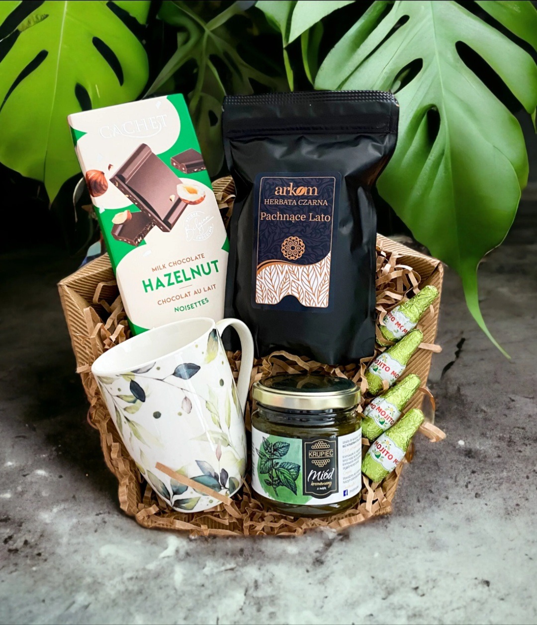 Kosz upominkowy z Dąbrowy Górniczej zawierający czarną herbatę, miód z dodatkiem mięty, czekoladę z orzechami laskowymi, ceramiczny kubek w liście oraz czekoladki w kształcie butelek mojito...