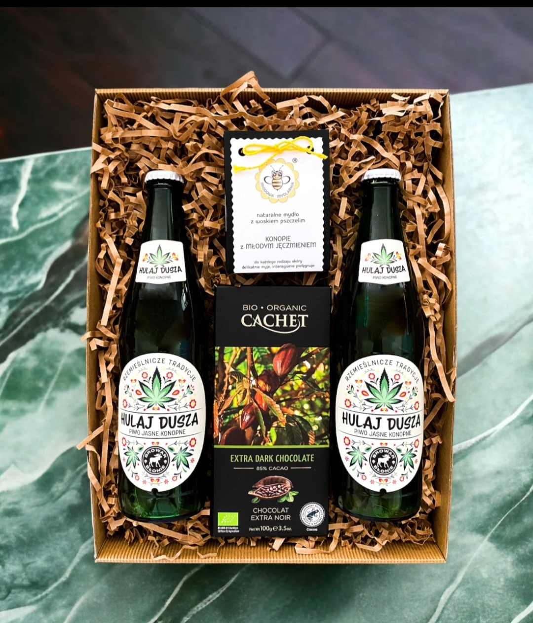 Upominkowy zestaw w kartonowym pudełku wypełnionym papierowym siankiem, zawierający dwie butelki piwa konopnego HULAJ DUSZA, tabliczkę gorzkiej czekolady BIO ORGANIC CACHET i mydło naturalne...