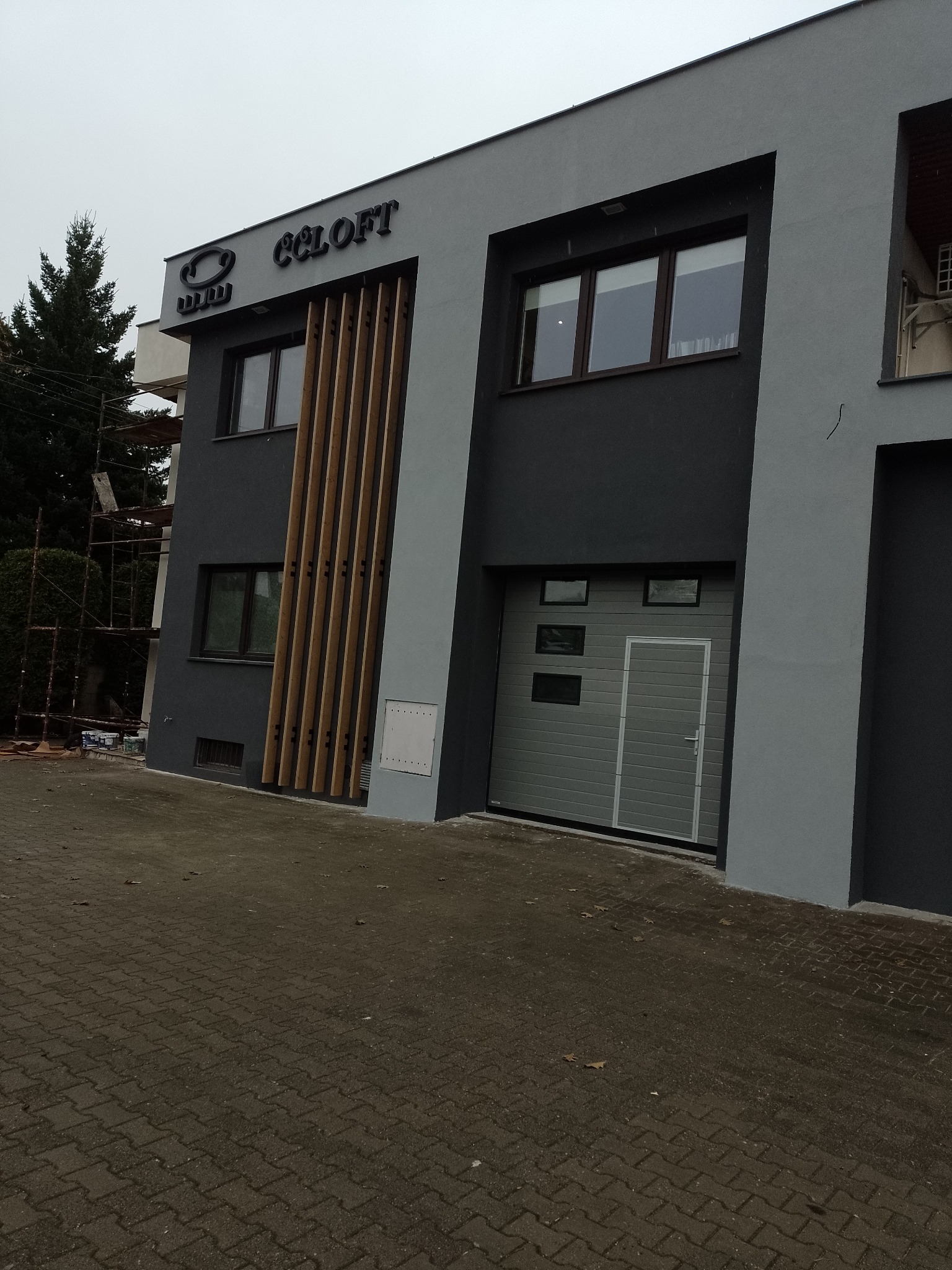 Dwukondygnacyjny budynek o szarej elewacji z drewnianymi pionowymi panelami dekoracyjnymi, widoczny szyld firmy CC LOFT i brama garażowa, częściowo zakryty rusztowaniami po lewej stronie.