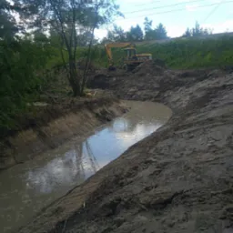 Wykopany rów wypełniony wodą w Tarnowie, z żółtą koparką CAT M315 w tle, sugeruje prace przy tworzeniu lub pogłębianiu zbiornika wodnego.