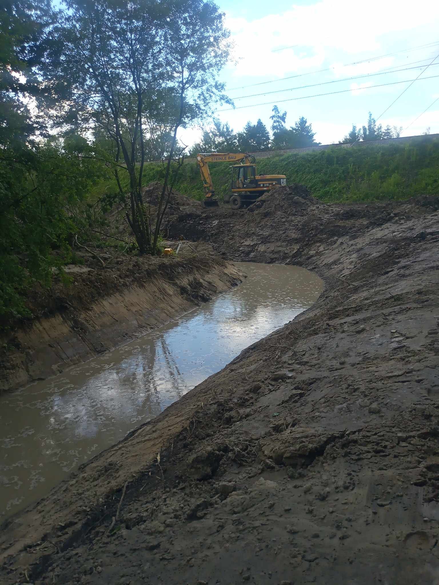Wykopany rów wypełniony wodą w Tarnowie, z żółtą koparką CAT M315 w tle, sugeruje prace przy tworzeniu lub pogłębianiu zbiornika wodnego.