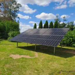 Panel słoneczny na metalowej konstrukcji ustawiony na trawniku w otoczeniu drzew i krzew&oacute;w, błękitne niebo z białymi chmurami w tle.