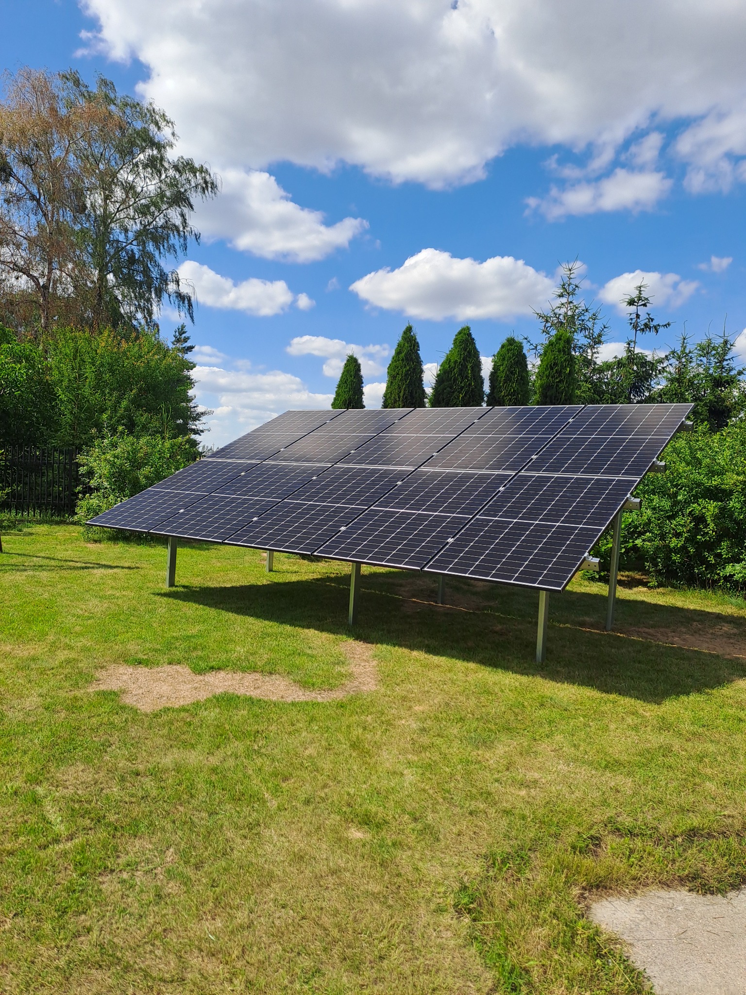 Panel słoneczny na metalowej konstrukcji ustawiony na trawniku w otoczeniu drzew i krzewów, błękitne niebo z białymi chmurami w tle.