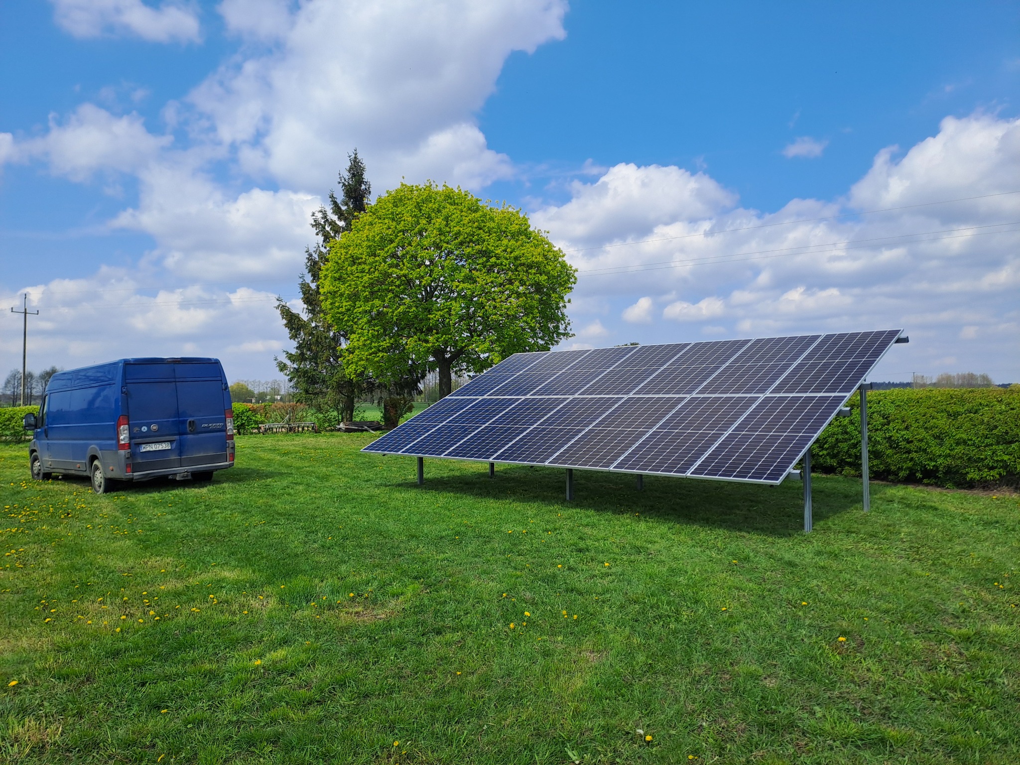 Panel słoneczny na metalowej konstrukcji, ustawiony na trawniku z widocznym niebieskim samochodem dostawczym i drzewem w tle, przy błękitnym niebie z chmurami.