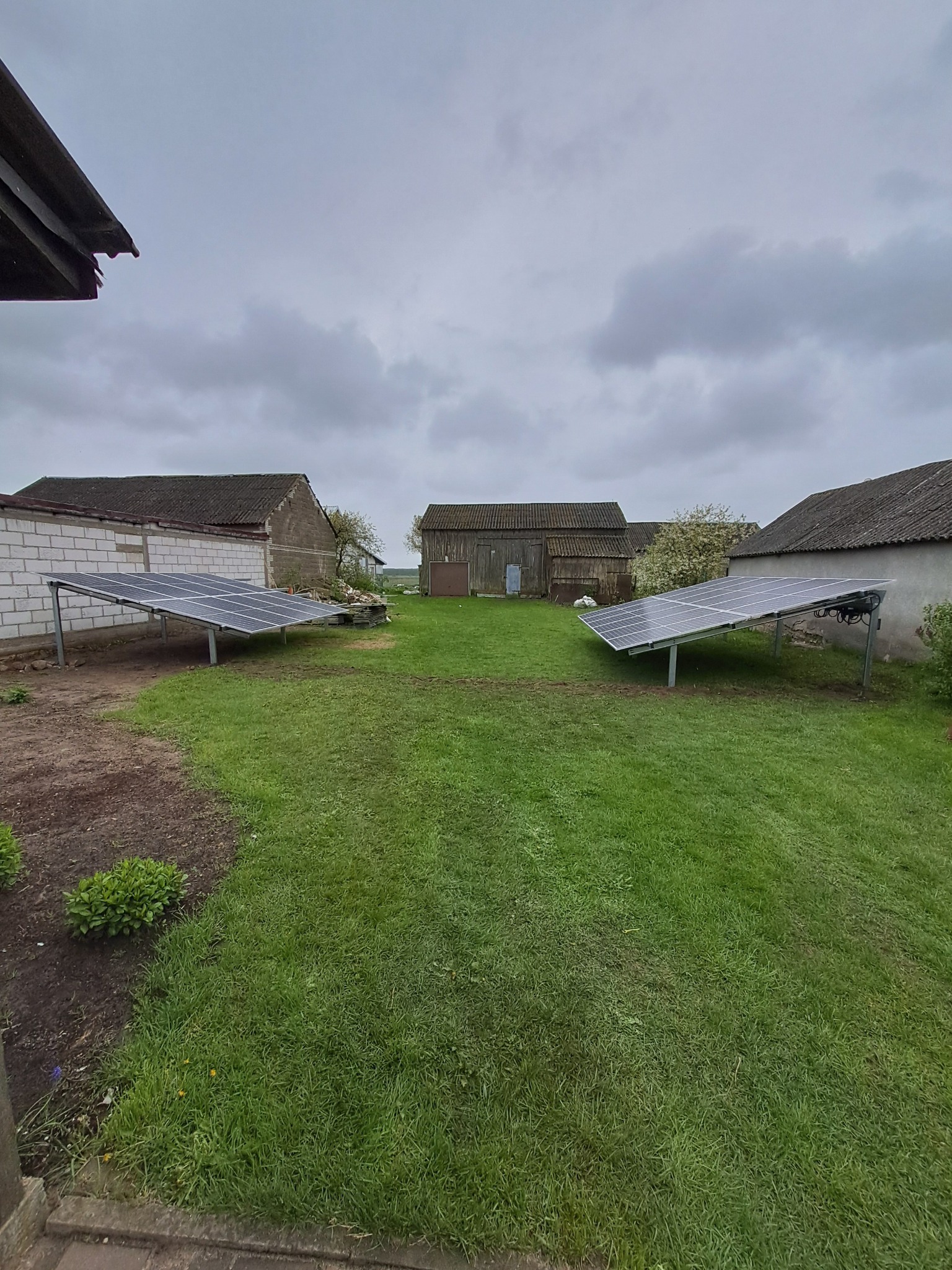 Dwa rzędy paneli słonecznych na metalowych stelażach, ustawione na trawiastym podwórku wiejskiego gospodarstwa w pochmurny dzień.