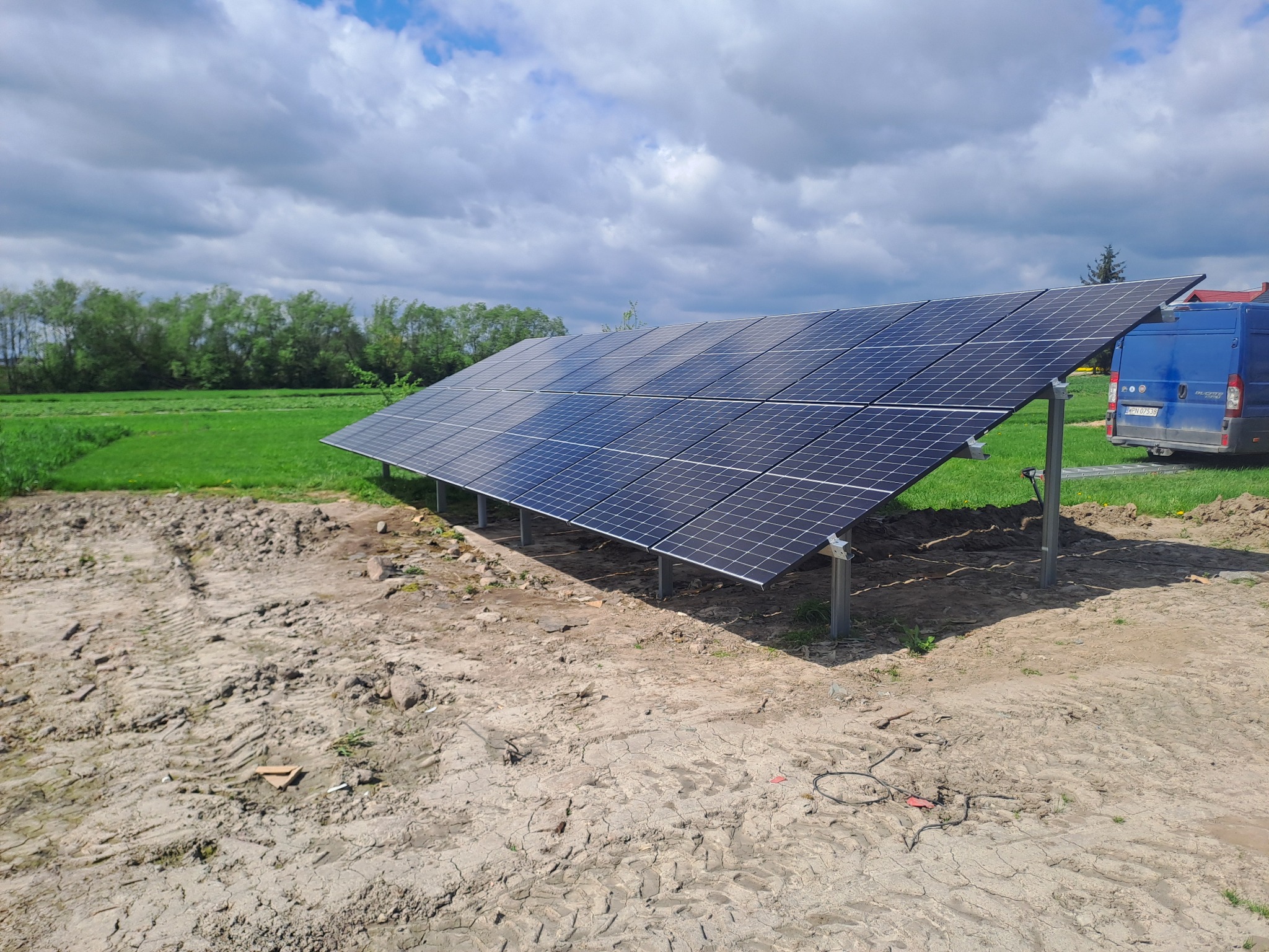 Instalacja paneli słonecznych na metalowej konstrukcji, widoczny niebieski samochód dostawczy w tle, teren wokół w trakcie prac ziemnych.