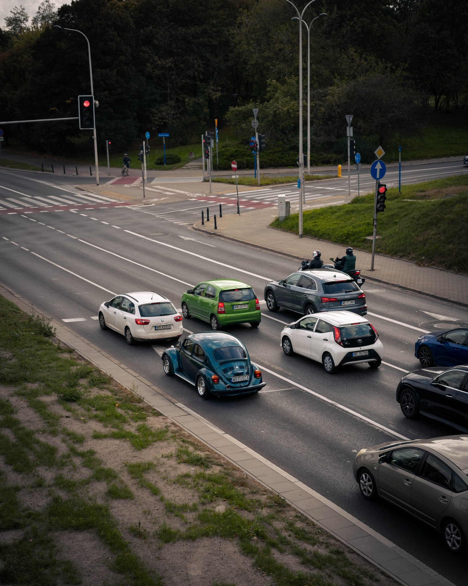 Turkusowy Volkswagen Garbus w korku miejskim, widziany z góry, z innymi samochodami i sygnalizacją świetlną w tle.