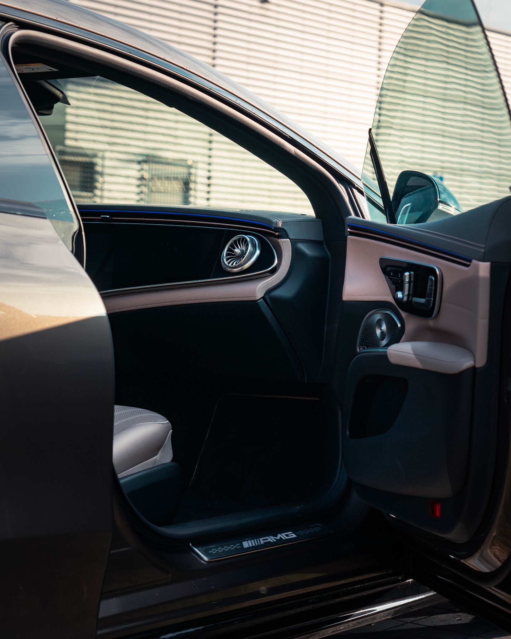 Wnętrze samochodu z otwartymi drzwiami, widoczny panel drzwiowy z elementami w kolorze beżowym i czarnym, srebrny głośnik i logo AMG na progu.