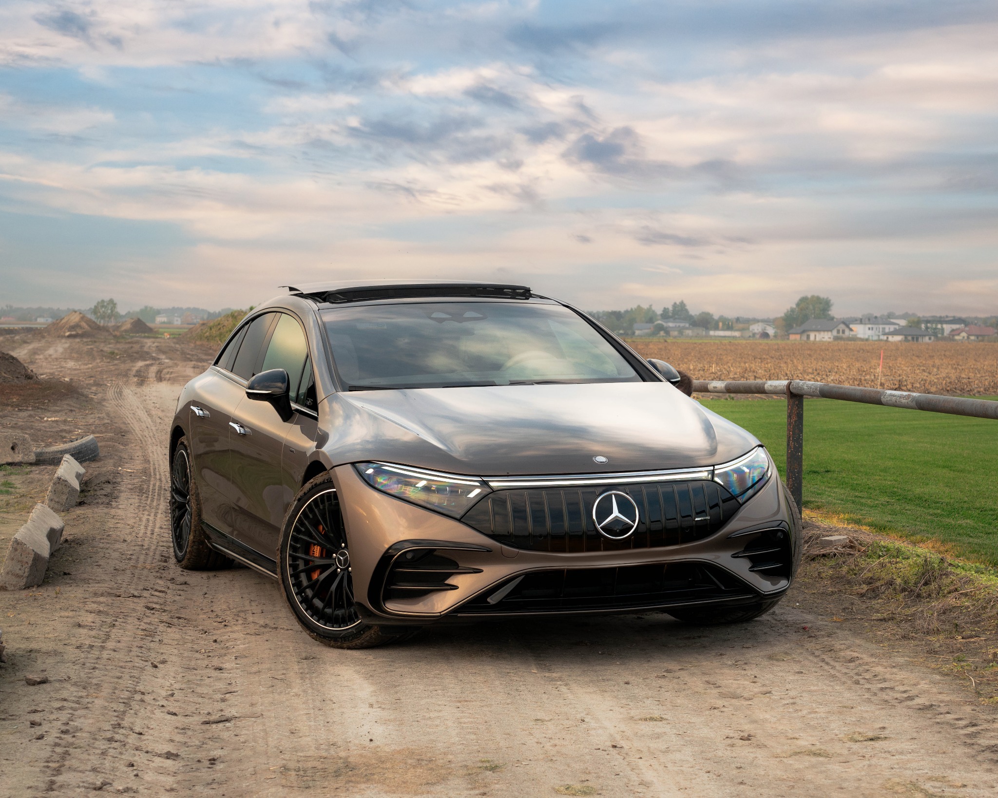 Elektryczny Mercedes EQS AMG w kolorze brązowym, sfotografowany w Legionowie na polnej drodze, z widocznym polem i zabudowaniami w tle, pod pochmurnym niebem.