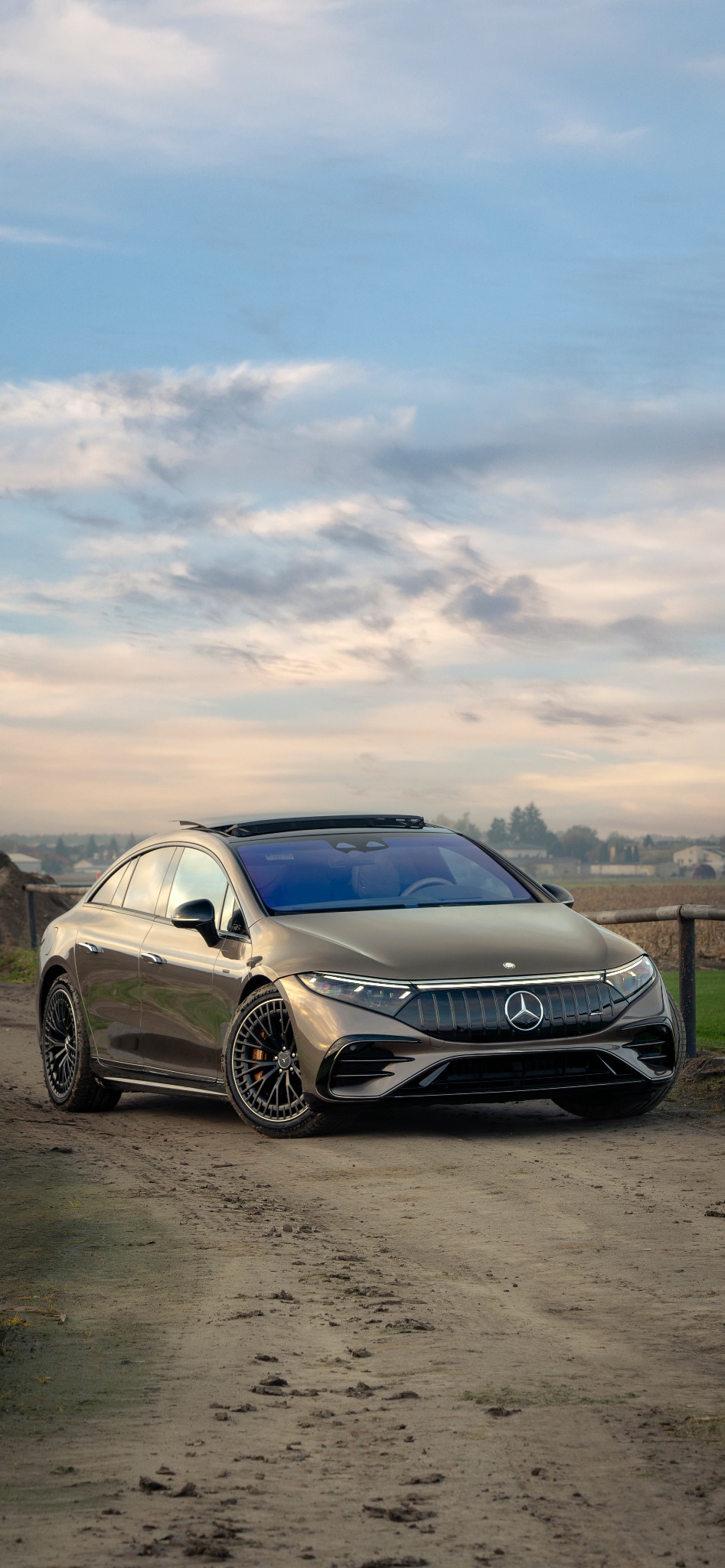 Elegancki, brązowy samochód elektryczny Mercedes-Benz EQS AMG na polnej drodze z widokiem na zachmurzone niebo i odległe zabudowania.