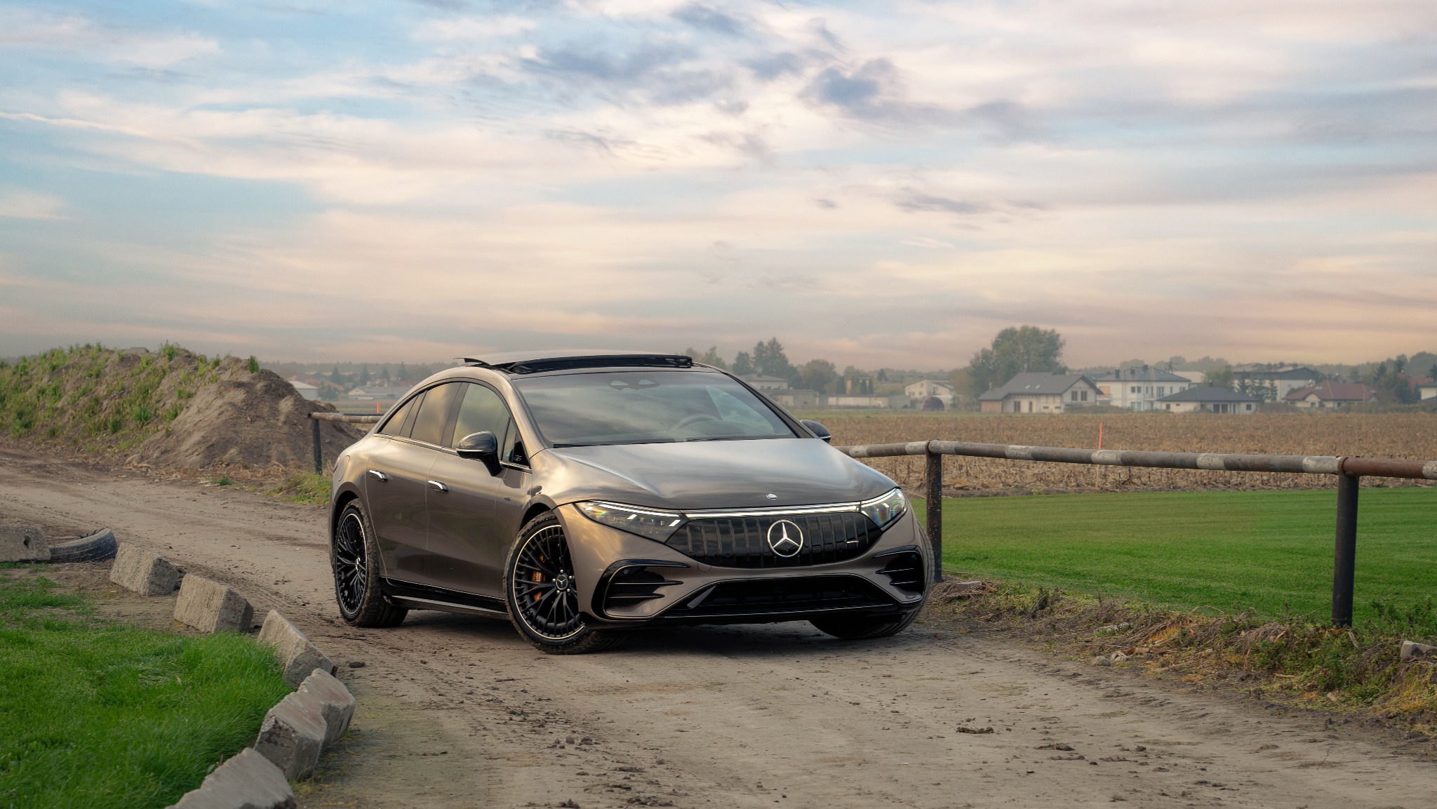 Szary elektryczny Mercedes-Benz EQS SUV z czarnymi felgami stoi na polnej drodze z widokiem na pola i domy w tle, oświetlony ciepłym światłem zachodzącego słońca.