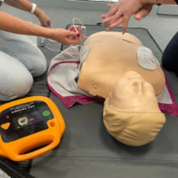 Instruktorzy podłączają elektrody do manekina podczas symulacji użycia defibrylatora AED Trainer; widoczny pomarańczowy defibrylator na szarym podłożu.