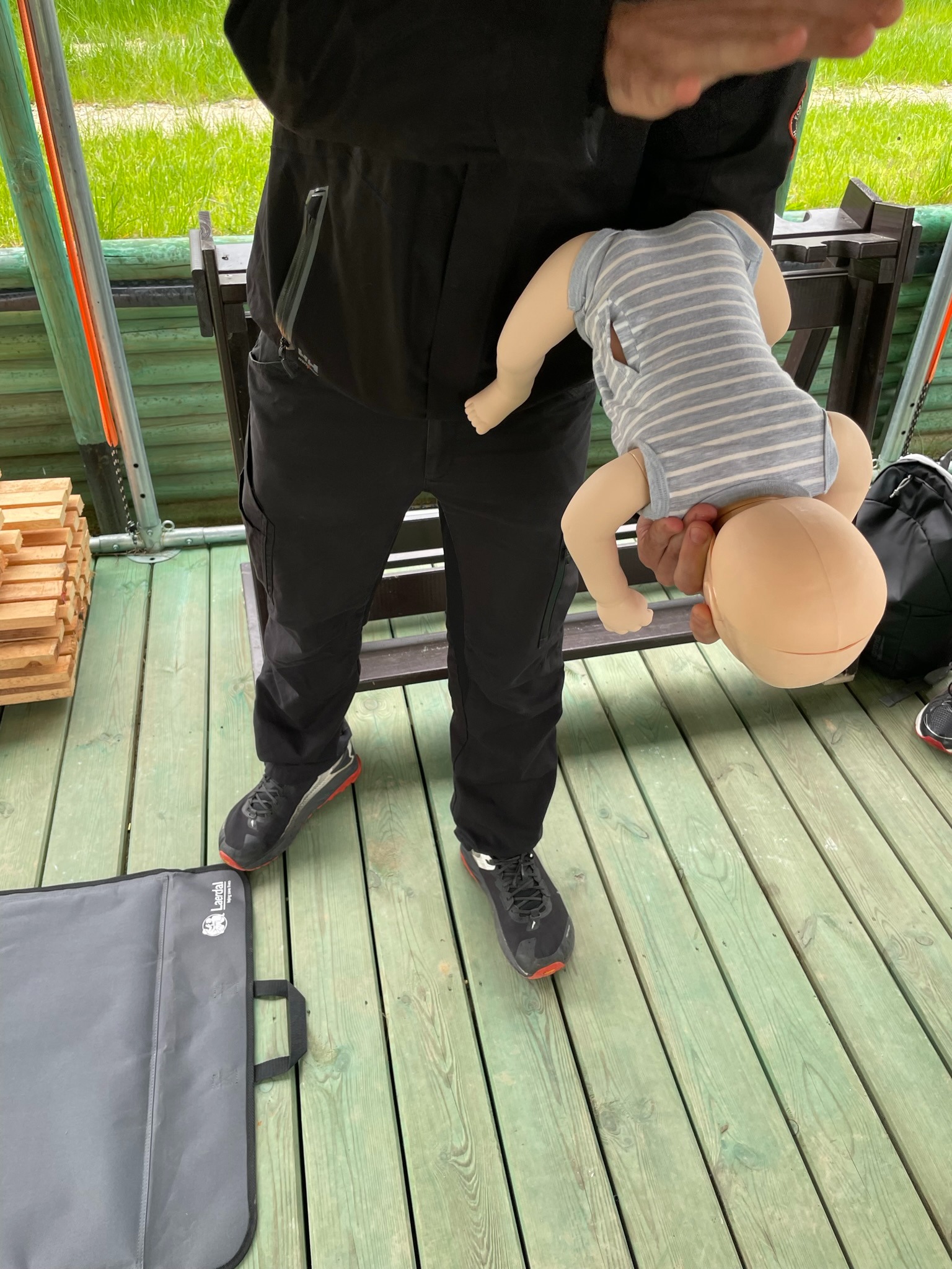 Instruktor trzymający manekina niemowlęcia do demonstracji, widoczny na tle zielonej trawy i drewnianej podłogi.