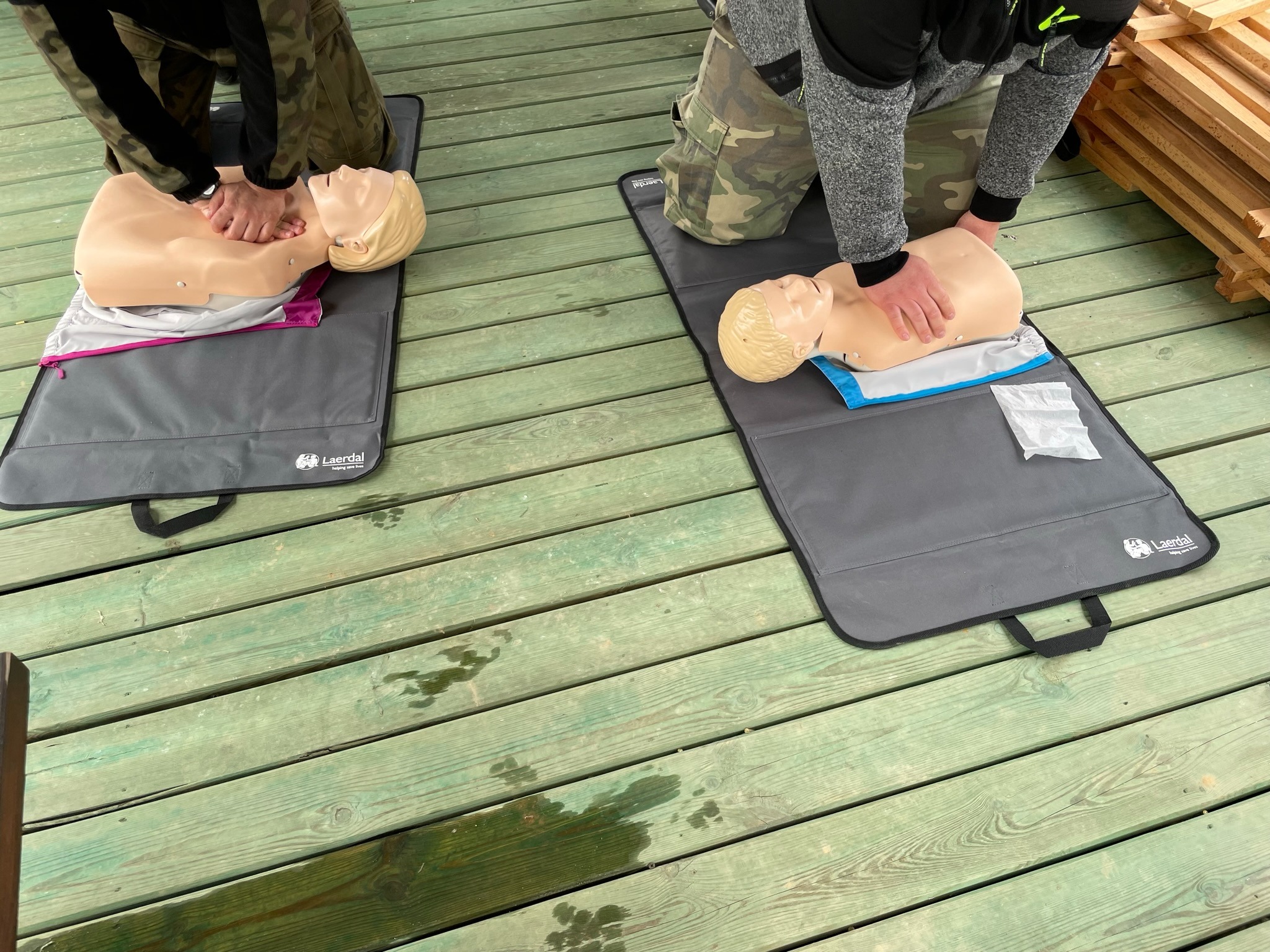 Dwie osoby ćwiczą resuscytację krążeniowo-oddechową na manekinach na zewnątrz, na zielonej, drewnianej podłodze, widoczny sprzęt Laerdal.