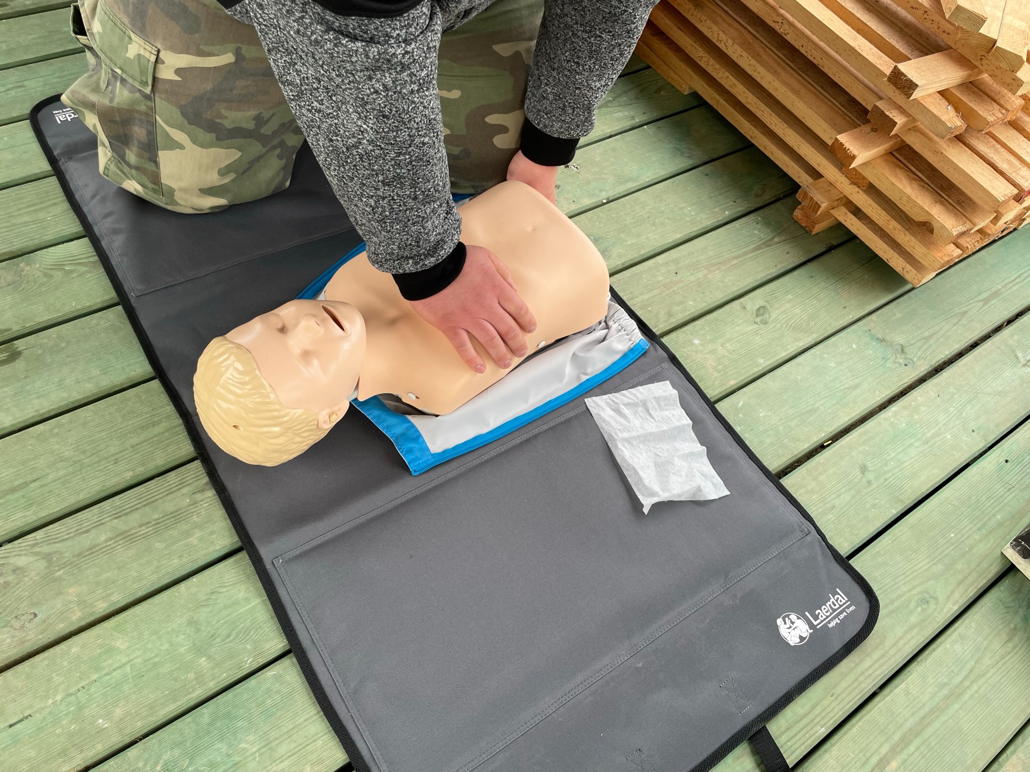 Instruktor demonstruje uciski klatki piersiowej na manekinie resuscytacyjnym Laerdal na tle zielonej drewnianej podłogi i stosu drewna.
