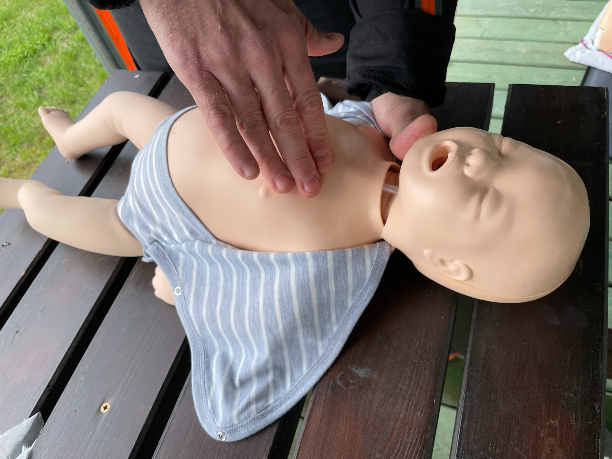 Instruktor demonstruje uciskanie klatki piersiowej na manekinie niemowlęcia podczas zajęć z pierwszej pomocy; manekin ubrany w body w paski leży na drewnianym stole